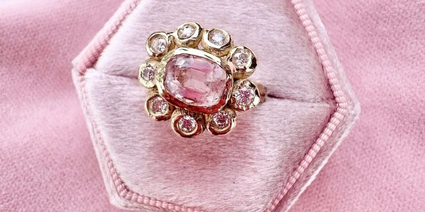 Ring with a pink gemstone and gold accents on a hexagonal pink velvet cushion.
