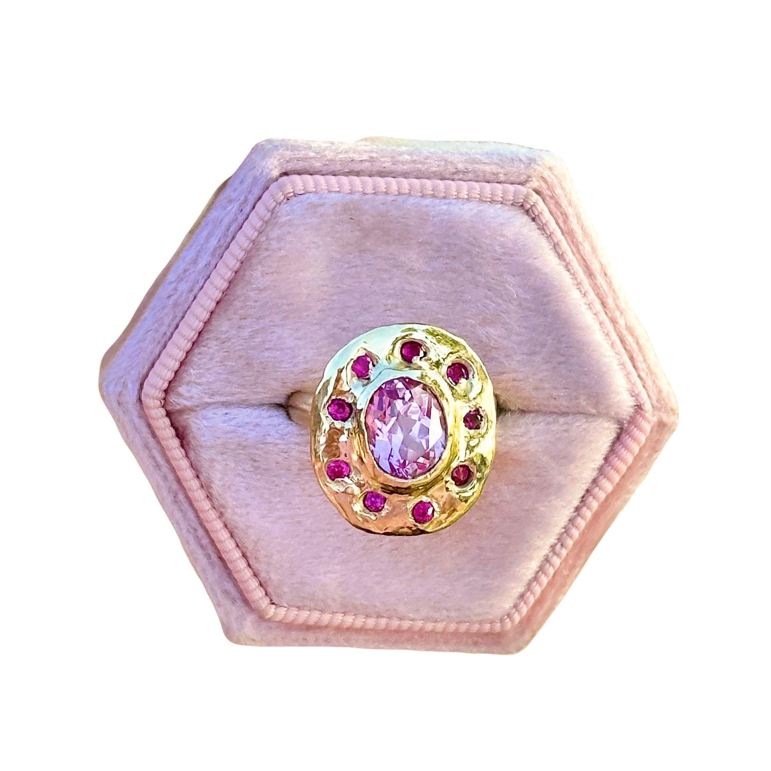 Ring with a pink gemstone in a gold setting on a hexagonal pink velvet cushion.